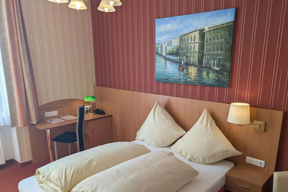 A hotel room with a bed, two pillows, a desk with a lamp, a chair, a nightstand with a lamp, and a painting of a canal scene on the wall.