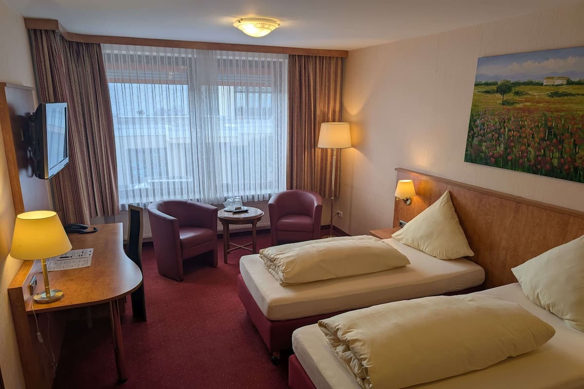 A hotel room with two beds, a desk with a lamp, a chair, a table, and a painting on the wall.