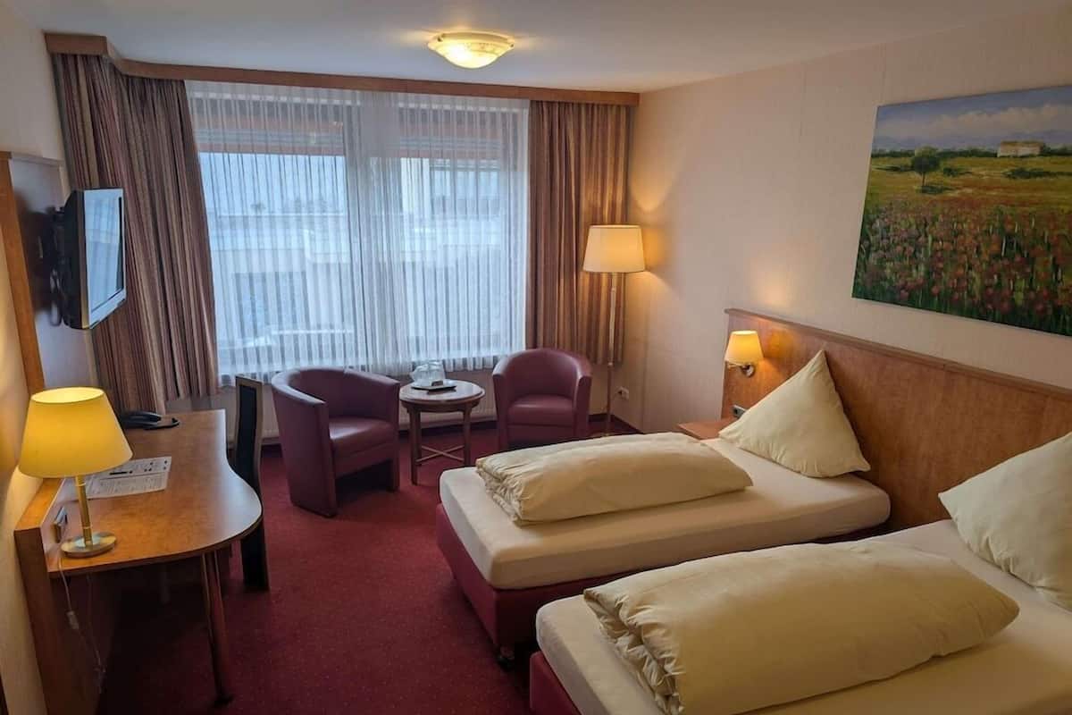 A hotel room with two beds, a desk with a lamp, a chair, a small table, and a painting on the wall.