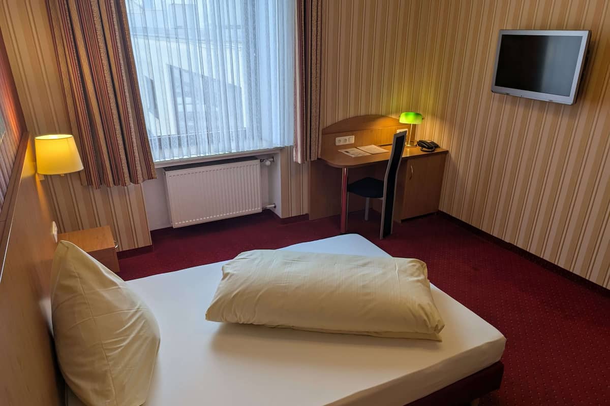 A hotel room with a single bed, a desk with a chair, a television mounted on the wall, and a window with curtains.