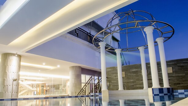 A spacious indoor swimming pool area with a modern design, featuring a central column structure and a staircase leading to an upper level. - Tara Hotel Yogyakarta (Yogyakarta)