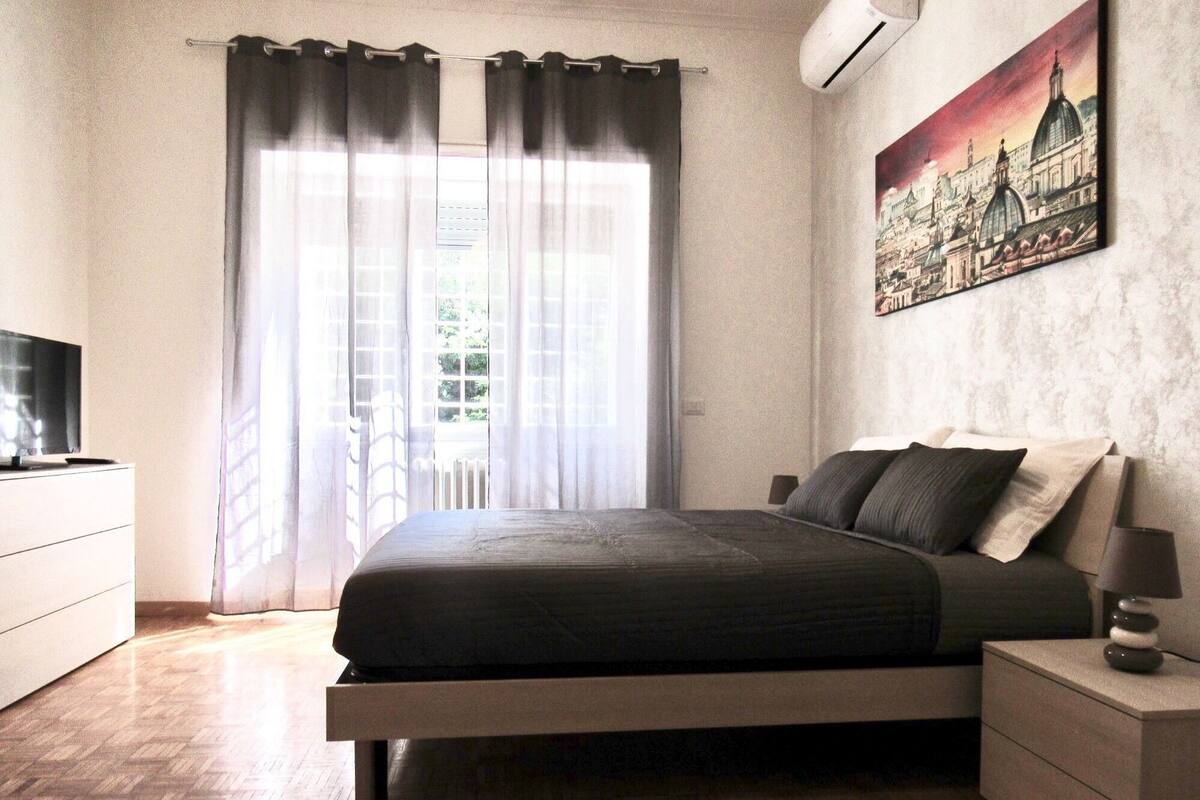 A bedroom with a bed, a TV, a painting, and a window with curtains.
