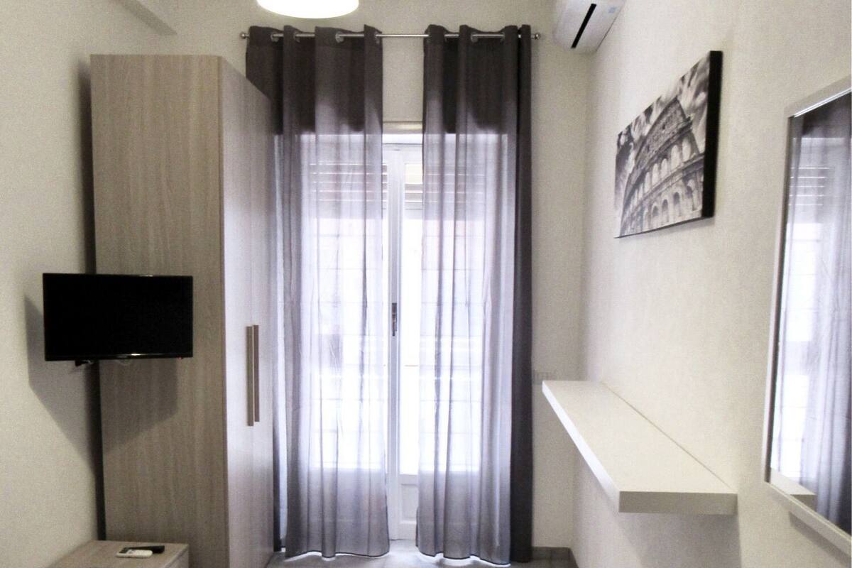 A small, modern bedroom with a bed, a TV mounted on the wall, a window with curtains, and a picture of a building on the wall.