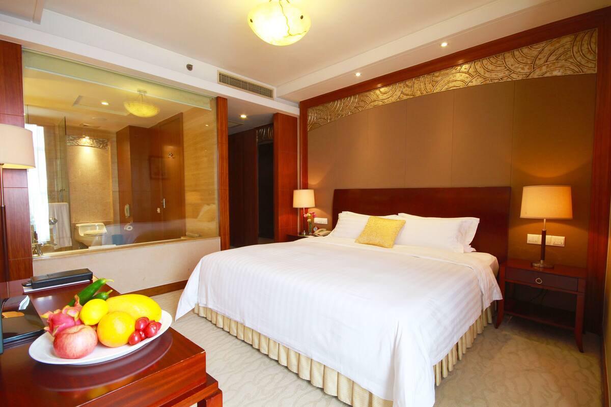 A hotel room with a large bed, two bedside lamps, a wooden nightstand, a mirror, and a plate of fruit on a wooden table.