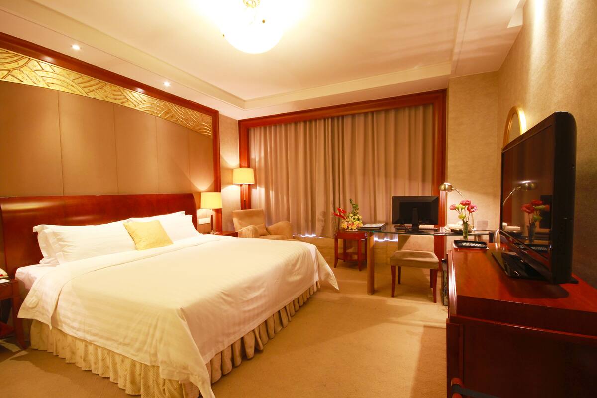 A hotel room with a large bed, a desk with a computer, a TV, and a piano.