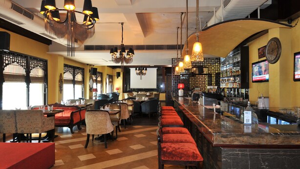 A bar area with a long counter, multiple seating options including bar stools and tables, and a projection screen in the background. - Qutub Residency (New Delhi)