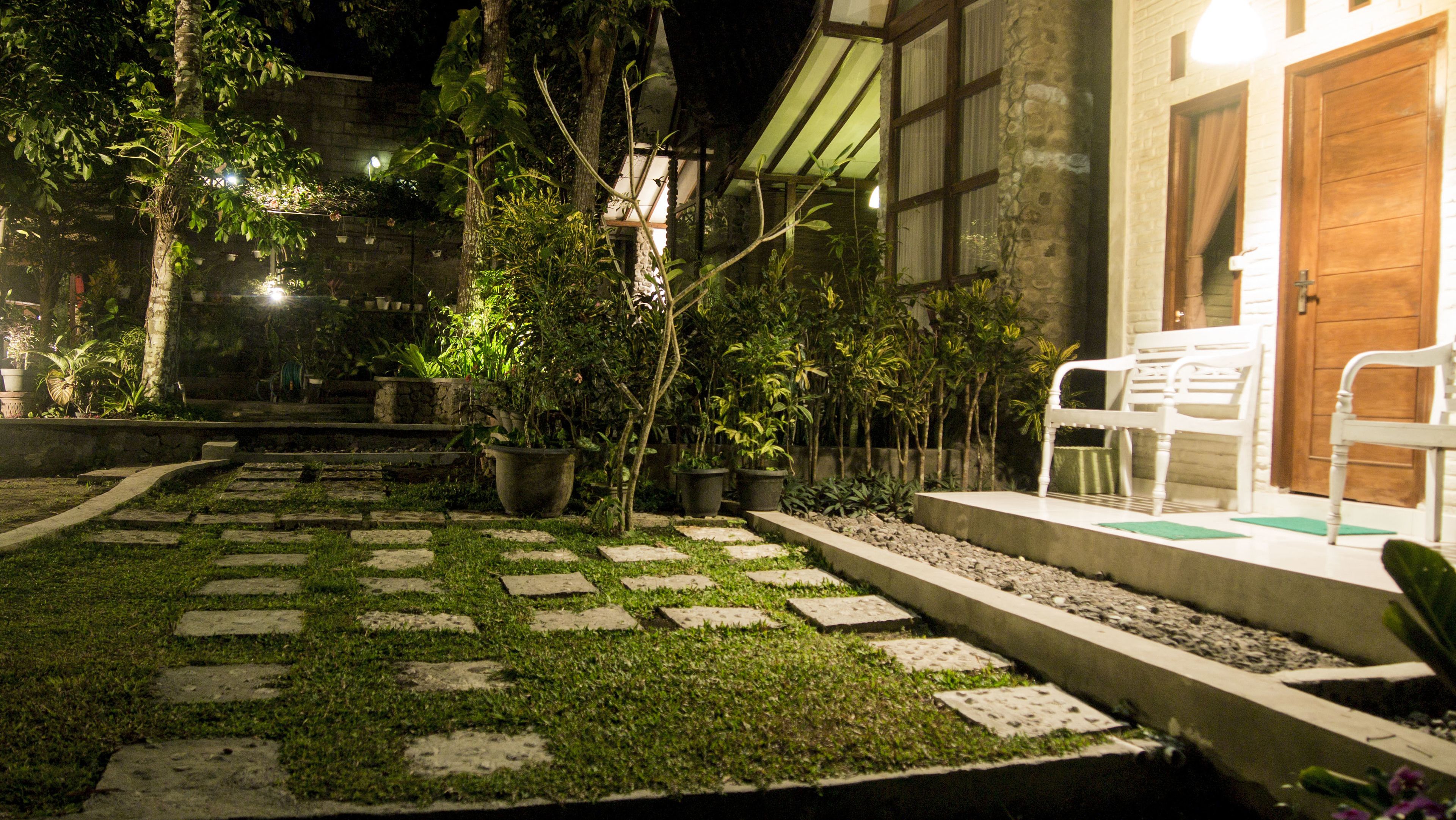 Un jardin nocturne avec des allées pavées de pierres, des plantes en pot et une chaise blanche près de l’entrée.