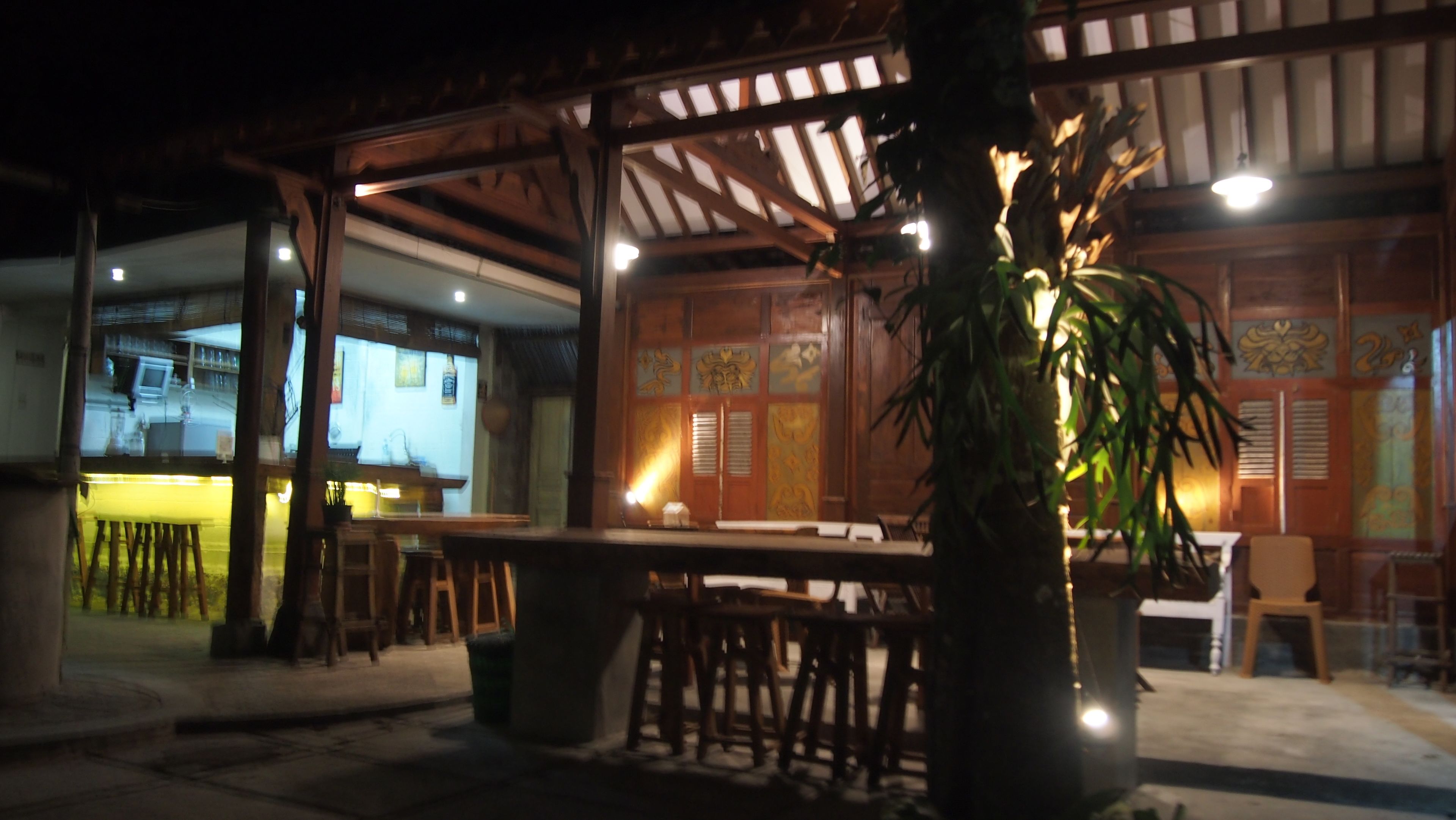 An outdoor dining area with wooden tables and chairs, a large potted plant, and a traditional wooden structure with decorative panels.