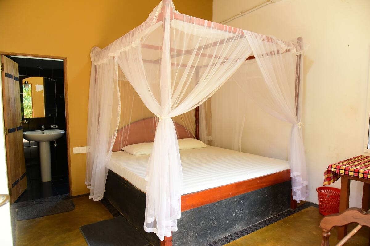 A room with a bed under a mosquito net, a sink, and a small table with a checkered tablecloth.