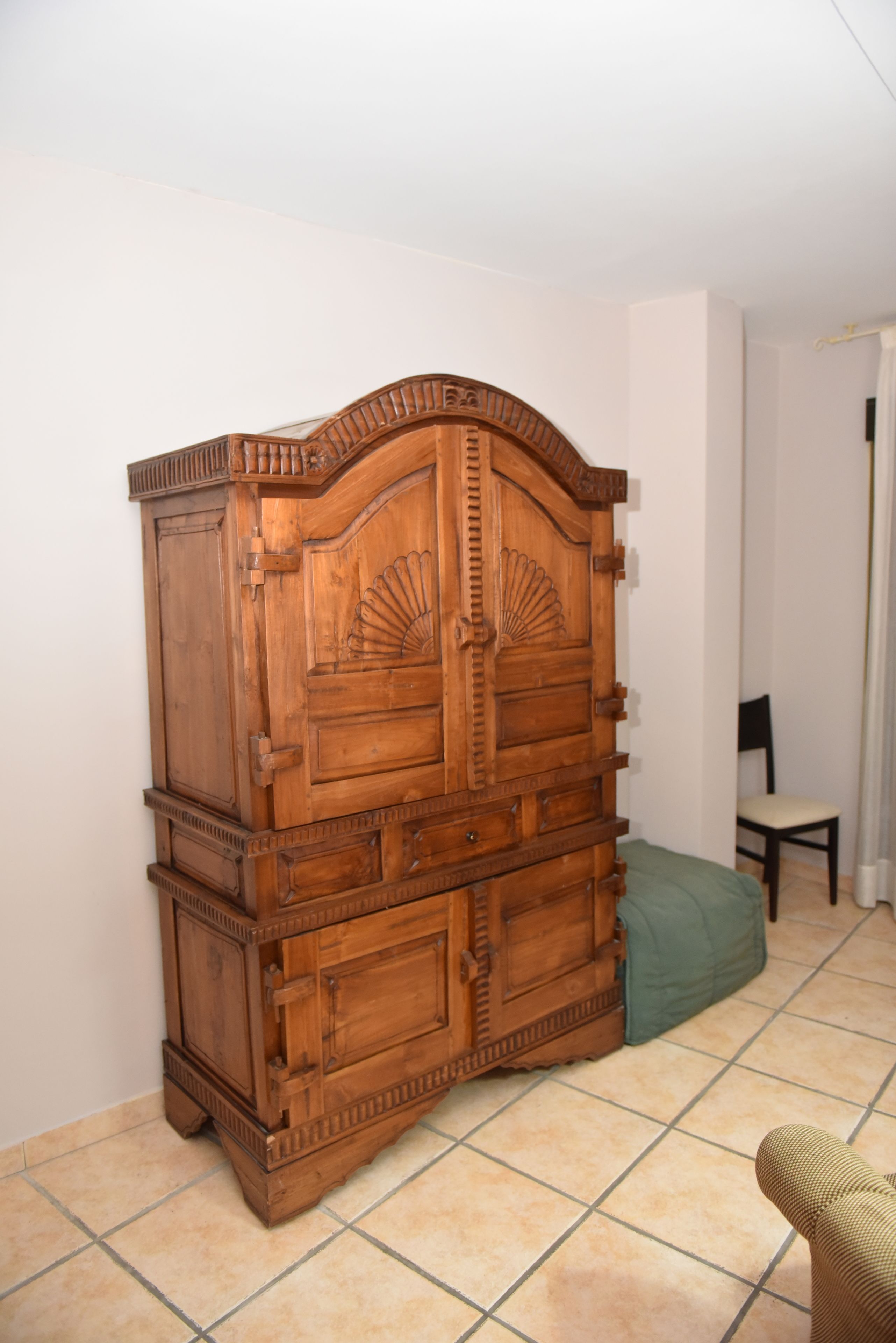 A wooden armoire with intricate carvings, a green cushion, and a chair in a room with tiled flooring.