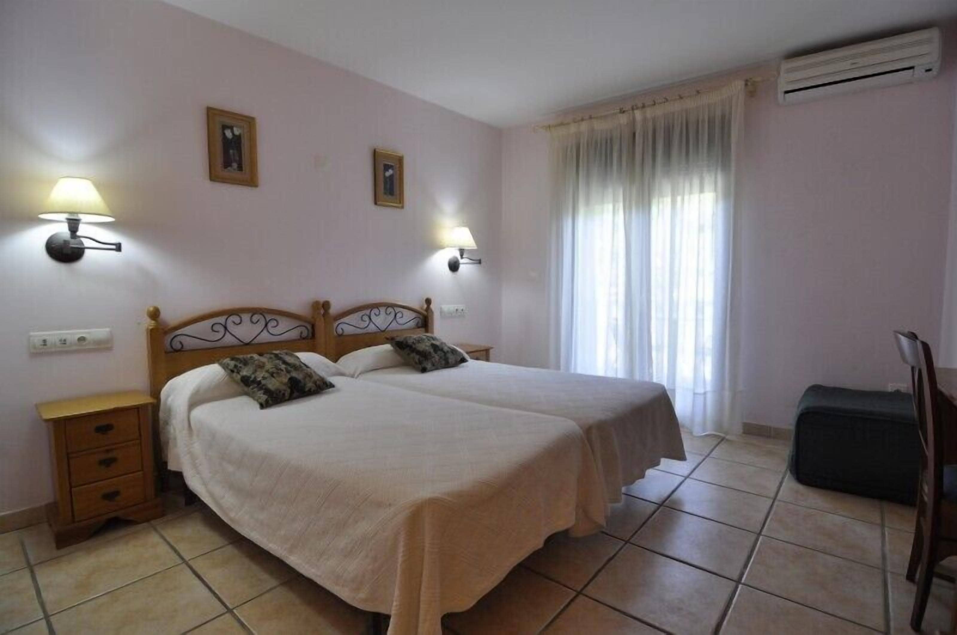 A double bed with two pillows, a bedside table with drawers, two wall-mounted lamps, and a window with curtains.