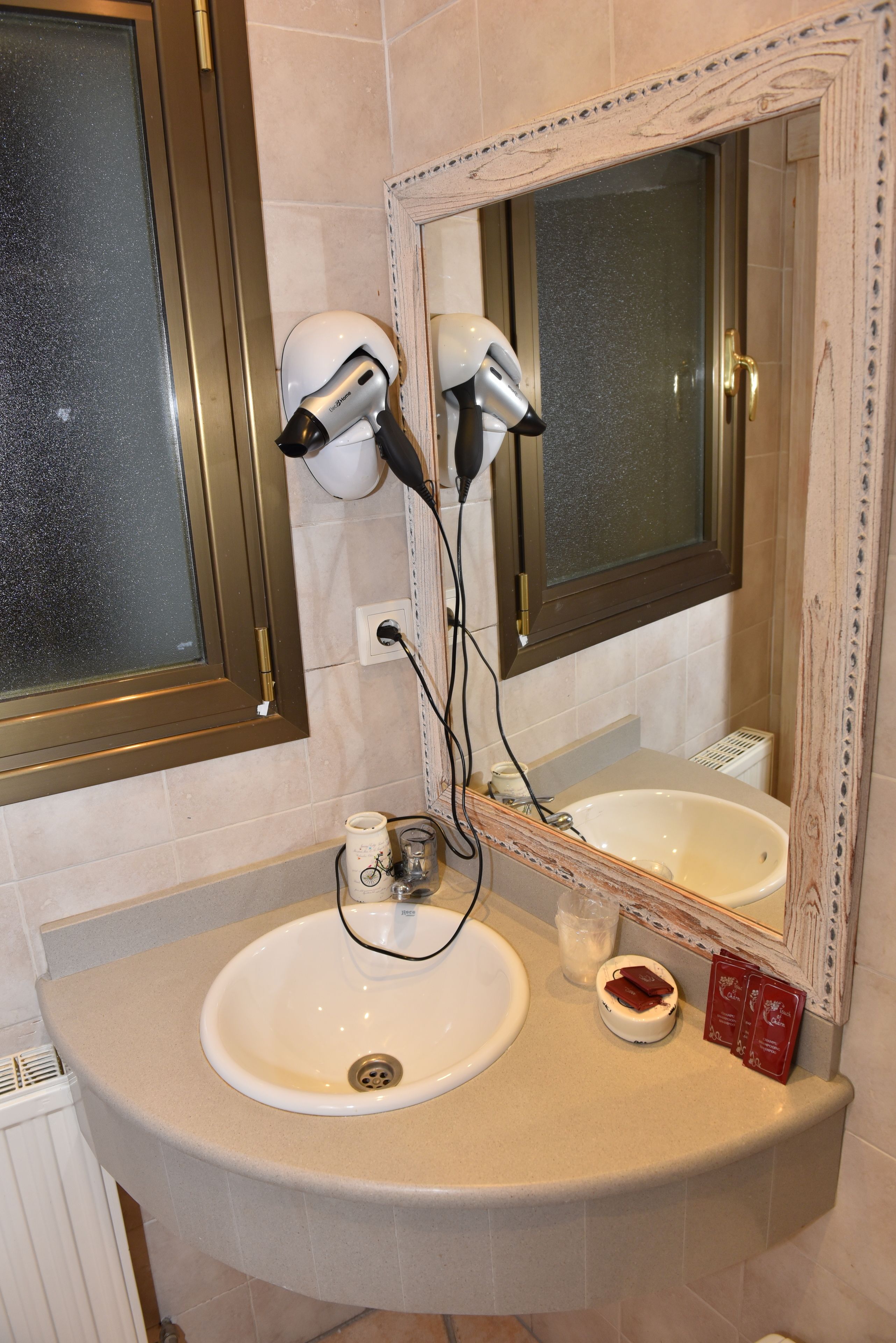 Une salle de bain avec un lavabo blanc, un miroir doté d’un cadre décoratif et deux sèche-cheveux fixés au mur.