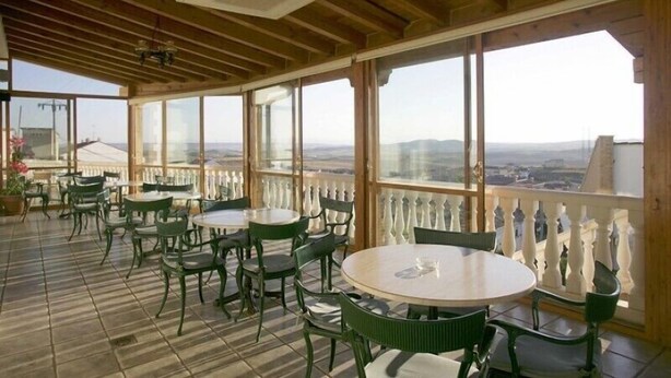 A covered outdoor seating area with tables and chairs, large windows, and a view of the landscape. - El Castillo (Larraga)
