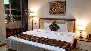 A hotel room with a large bed, two bedside lamps, a window with curtains, a wall-mounted air conditioner, and a painting above the bed. - Rene Hotel (Yogyakarta)