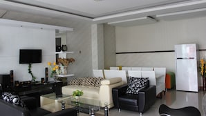 A modern living room with a flat-screen TV, a glass coffee table, a beige sofa, a black sofa, and a white refrigerator. - Rene Hotel (Yogyakarta)
