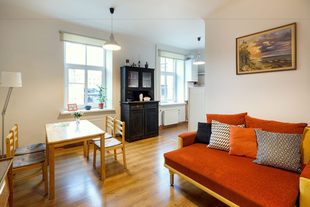A living room with a wooden floor, a sofa with patterned cushions, a dining table with chairs, and a cabinet.