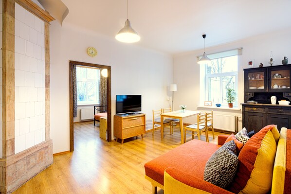 Una sala de estar moderna con piso de madera, un sofá con cojines estampados, una zona de comedor con mesa y sillas, un televisor y una cocina con alacenas y un ventanal.