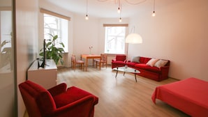 Una sala de estar moderna con un sofá y un sillón rojos, una mesa de centro de vidrio y una zona de comedor con una mesa y sillas de madera. Hay un ventanal grande que ofrece luz natural y vistas de edificios.