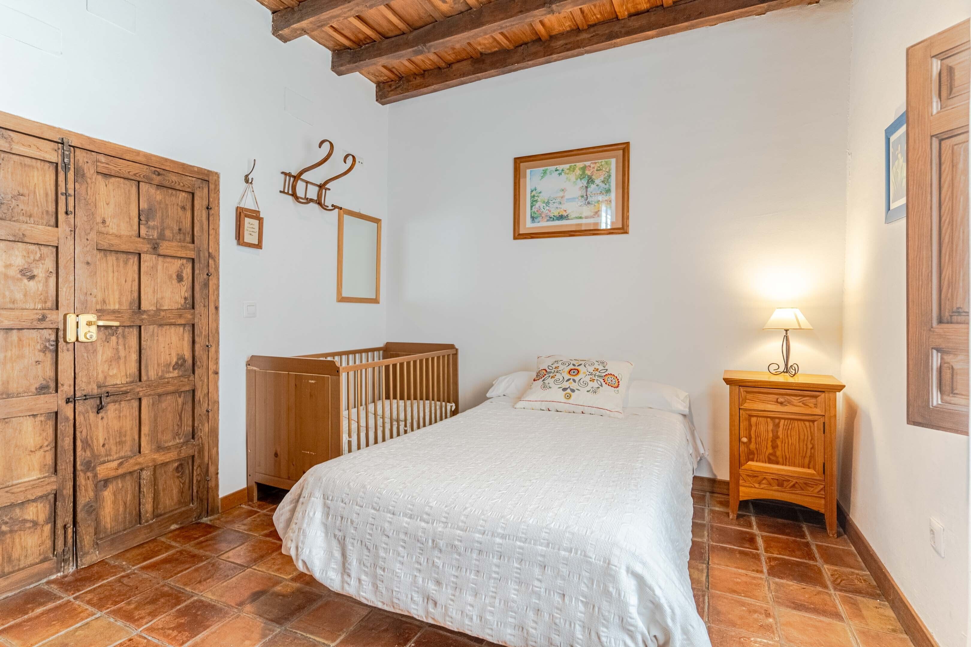 Un dormitorio con puerta de madera, una cuna, una cama con colcha blanca, una mesita de noche con lámpara y un cuadro enmarcado en la pared.