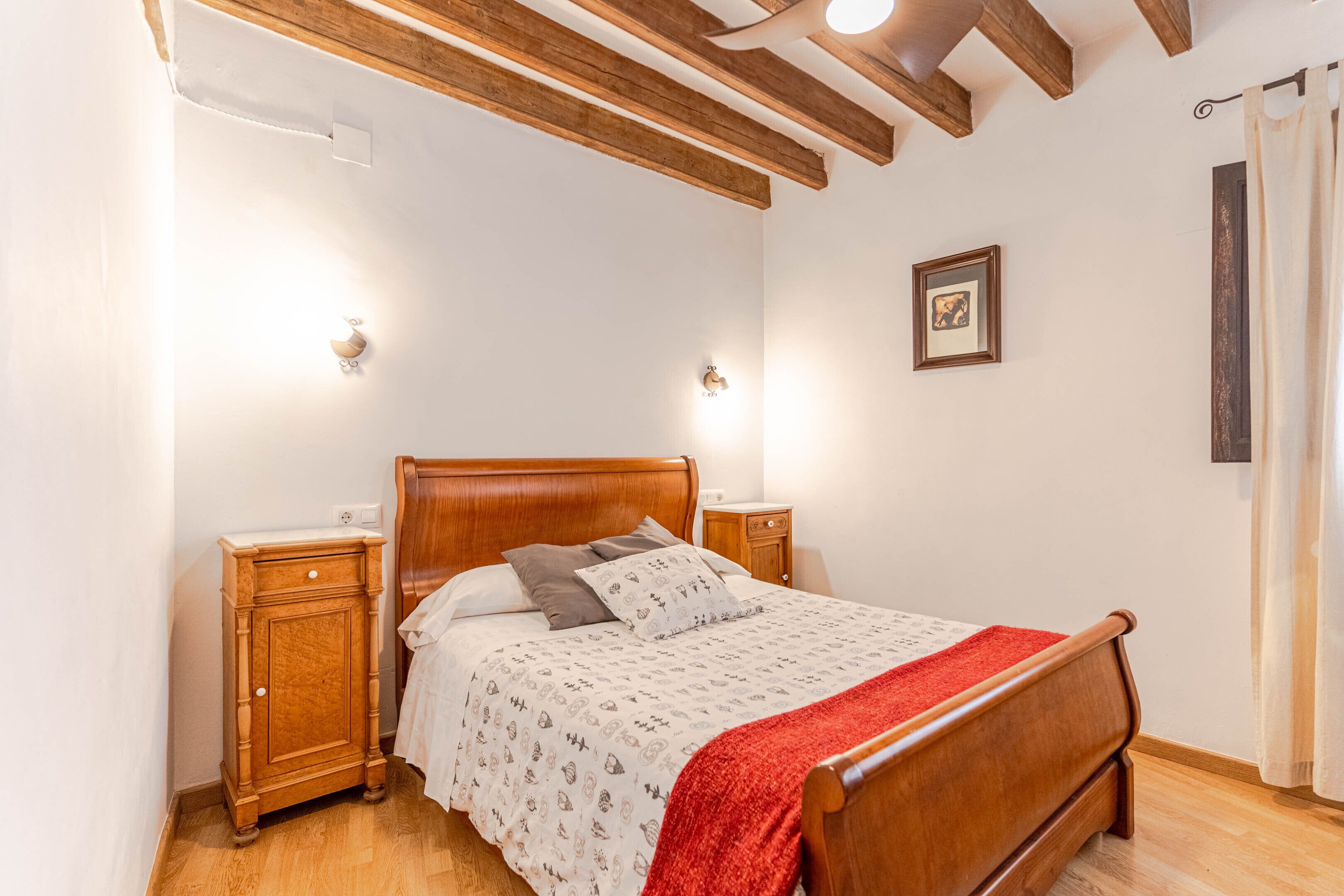 Un dormitori amb un llit de fusta, dues taules de nit, una tauleta auxiliar i un sostre amb bigues al descobert.