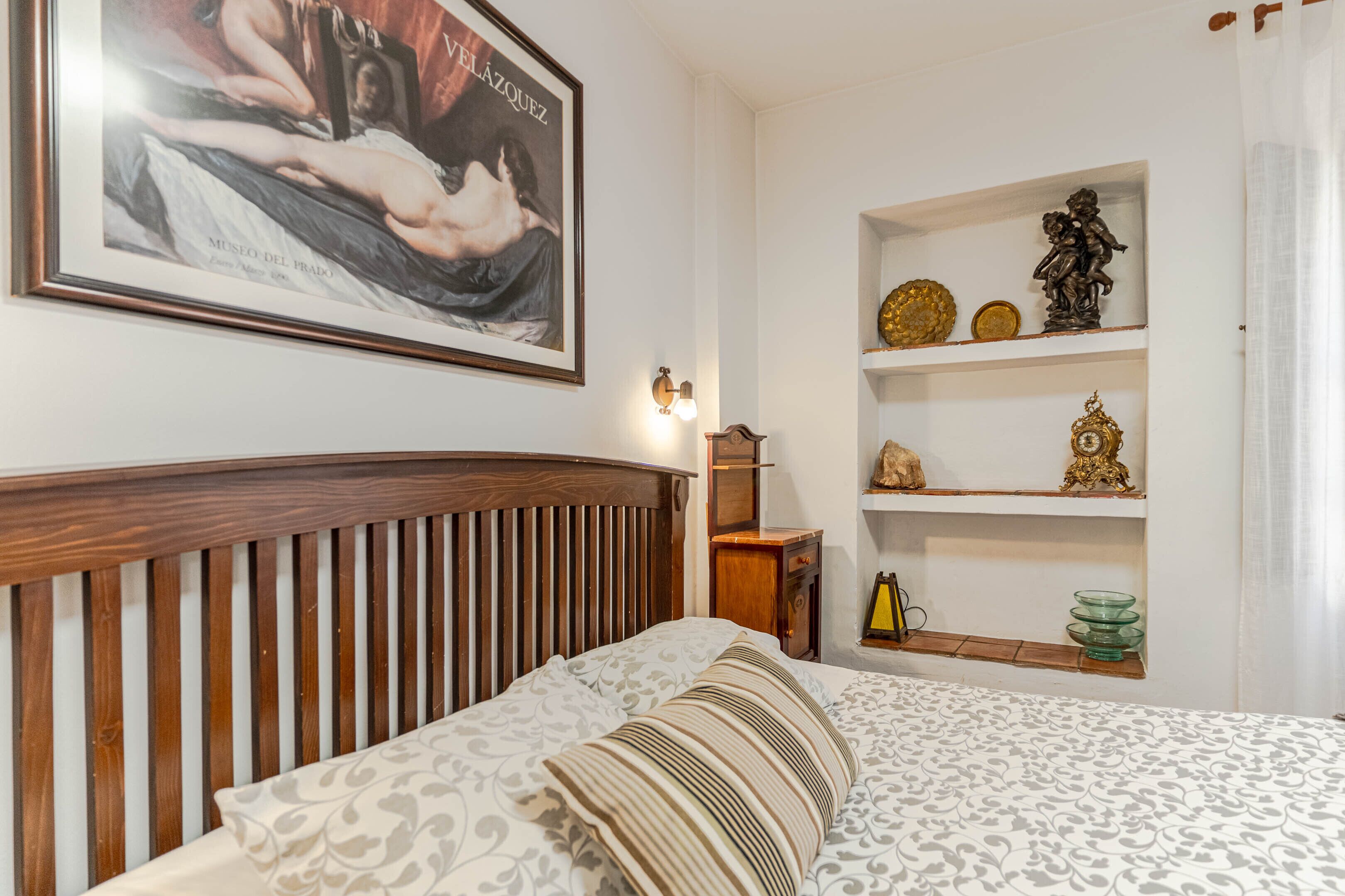 Un dormitorio con cabecera de madera, cama con ropa de cama estampada, estante en la pared con objetos decorativos y una obra enmarcada sobre la cama.