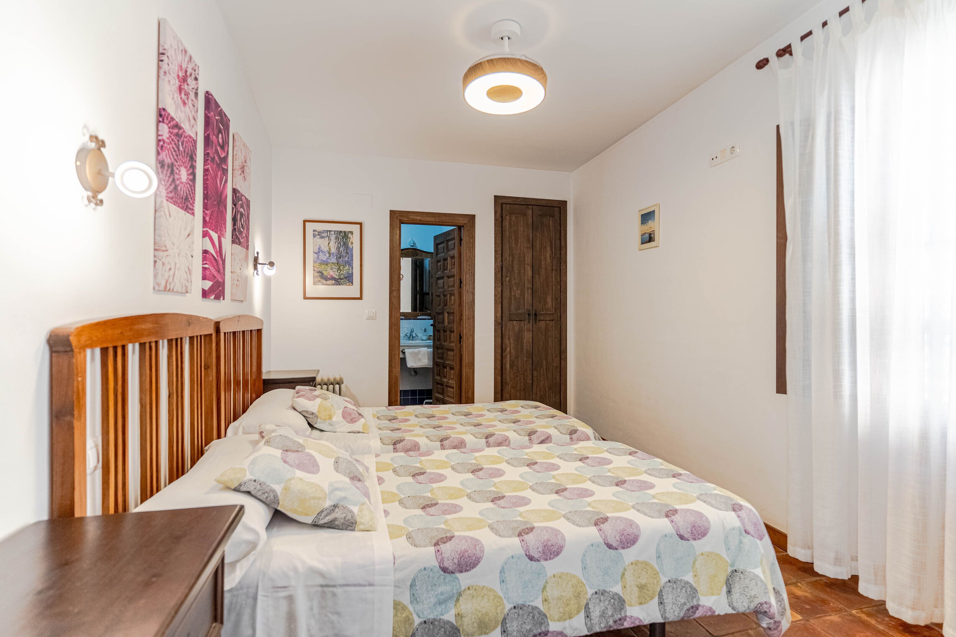 A bedroom with a wooden headboard, a bed with a multicolored circular pattern bedspread, a nightstand, and a wall-mounted lamp. There is a door leading to a bathroom and a hallway.