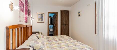 A bedroom with a wooden headboard, a bed with a multicolored circular pattern bedspread, a nightstand, and a wall-mounted lamp. There is a door leading to a bathroom and a hallway.