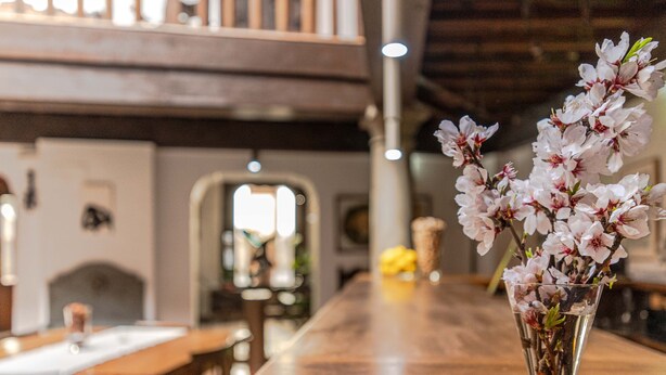 Een eethoek met een houten tafel en stoelen, een vaas met bloemen op de tafel, en uitzicht op het interieur met een hoge plafond en een trap.