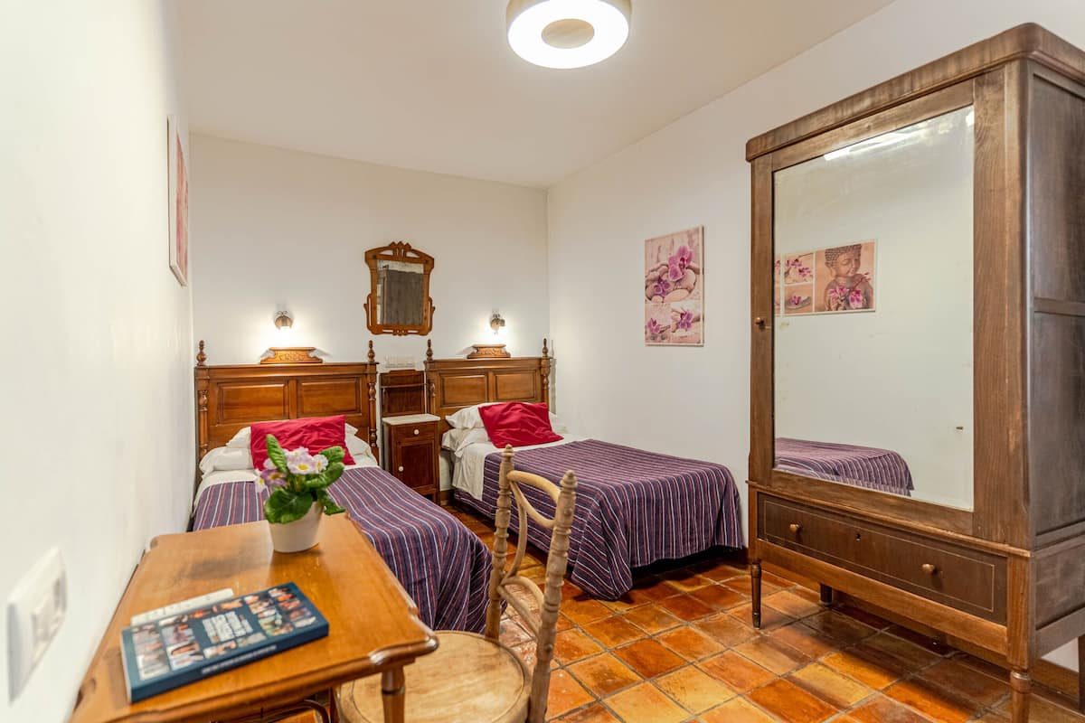Un dormitorio con dos camas, un armario de madera, un espejo, una mesita de noche con lámpara y una pequeña mesa con un libro.