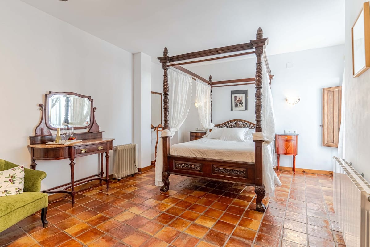 Una cama con dosel, una mesita de noche de madera, un sillón verde y un espejo en la pared.