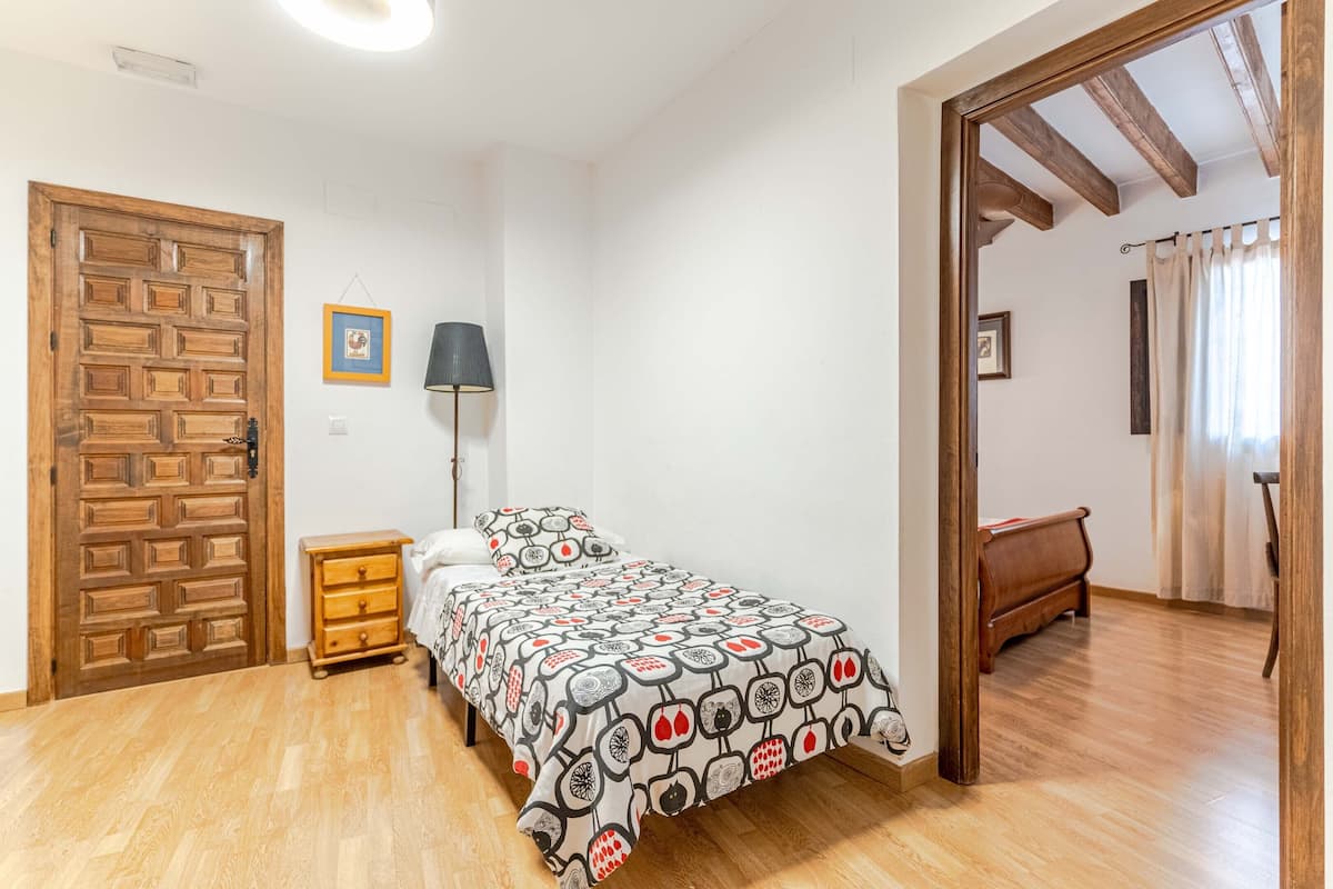 Un dormitorio con puerta de madera, cama con colcha estampada, mesita de noche y lámpara.