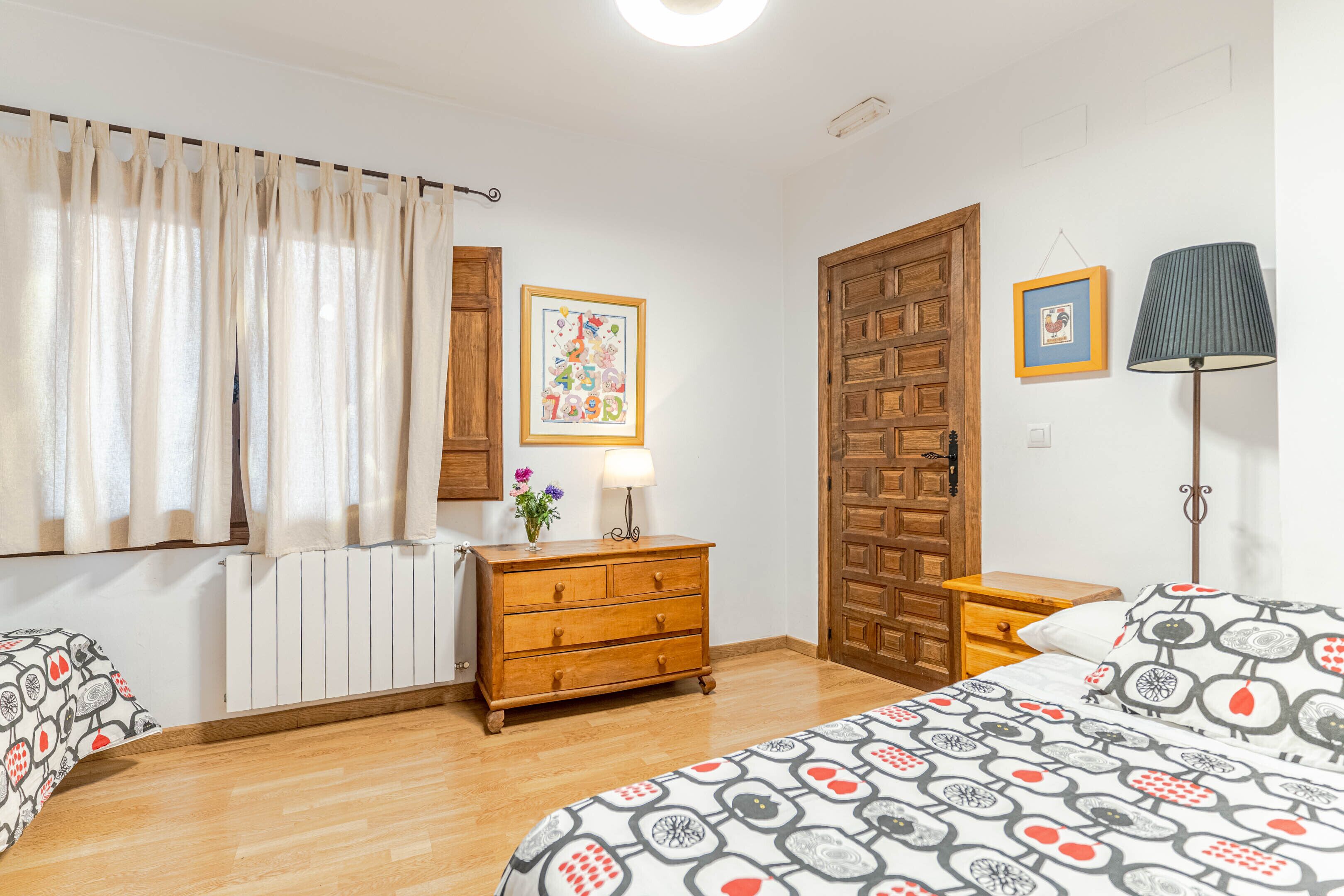 Un dormitorio con un aparador de madera, una cama con ropa de cama estampada, un radiador, una puerta de madera y un cuadro en la pared.