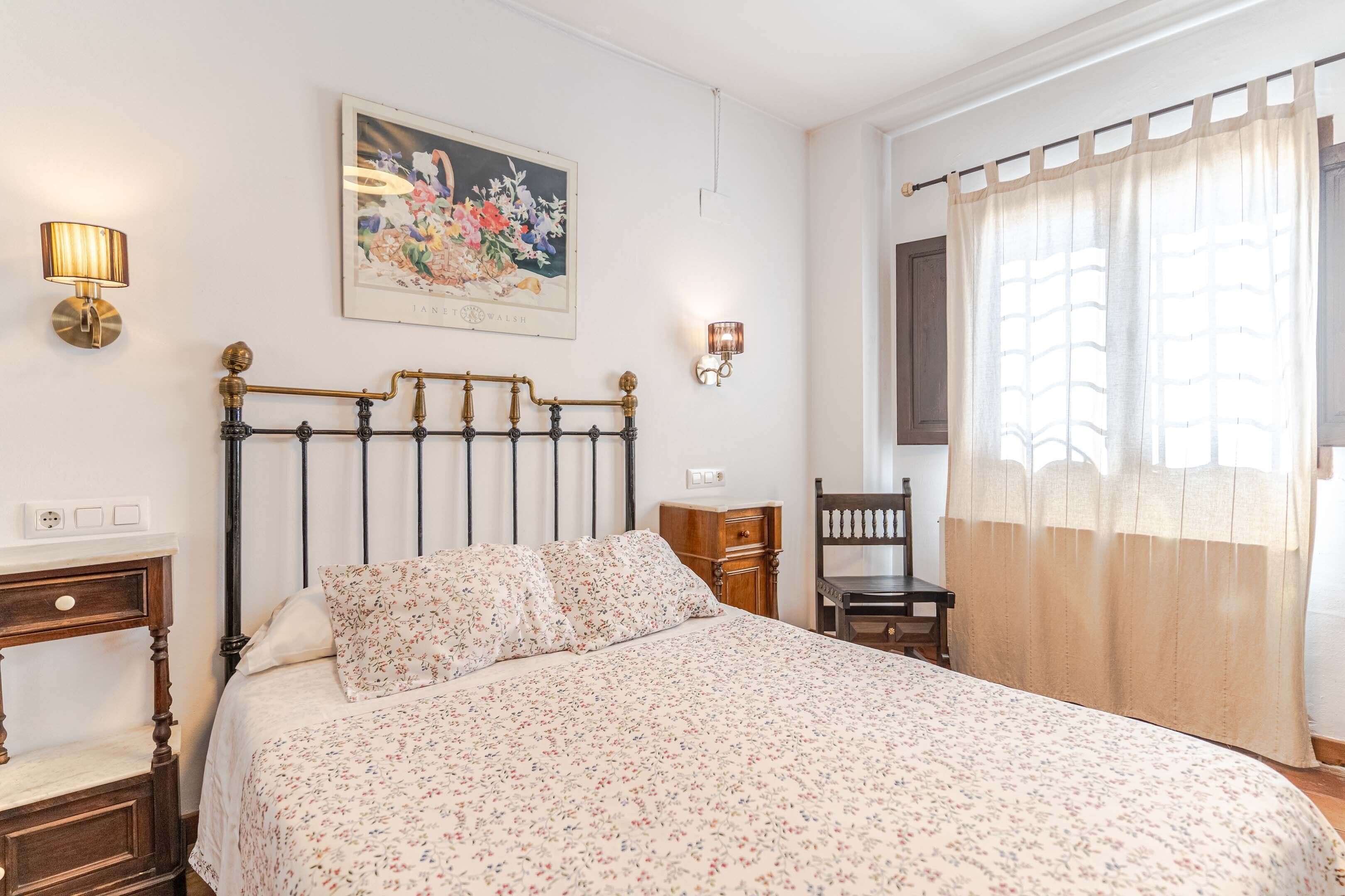 Un dormitorio con una cama, una mesita de noche, una silla, una lámpara de pared y un cuadro.