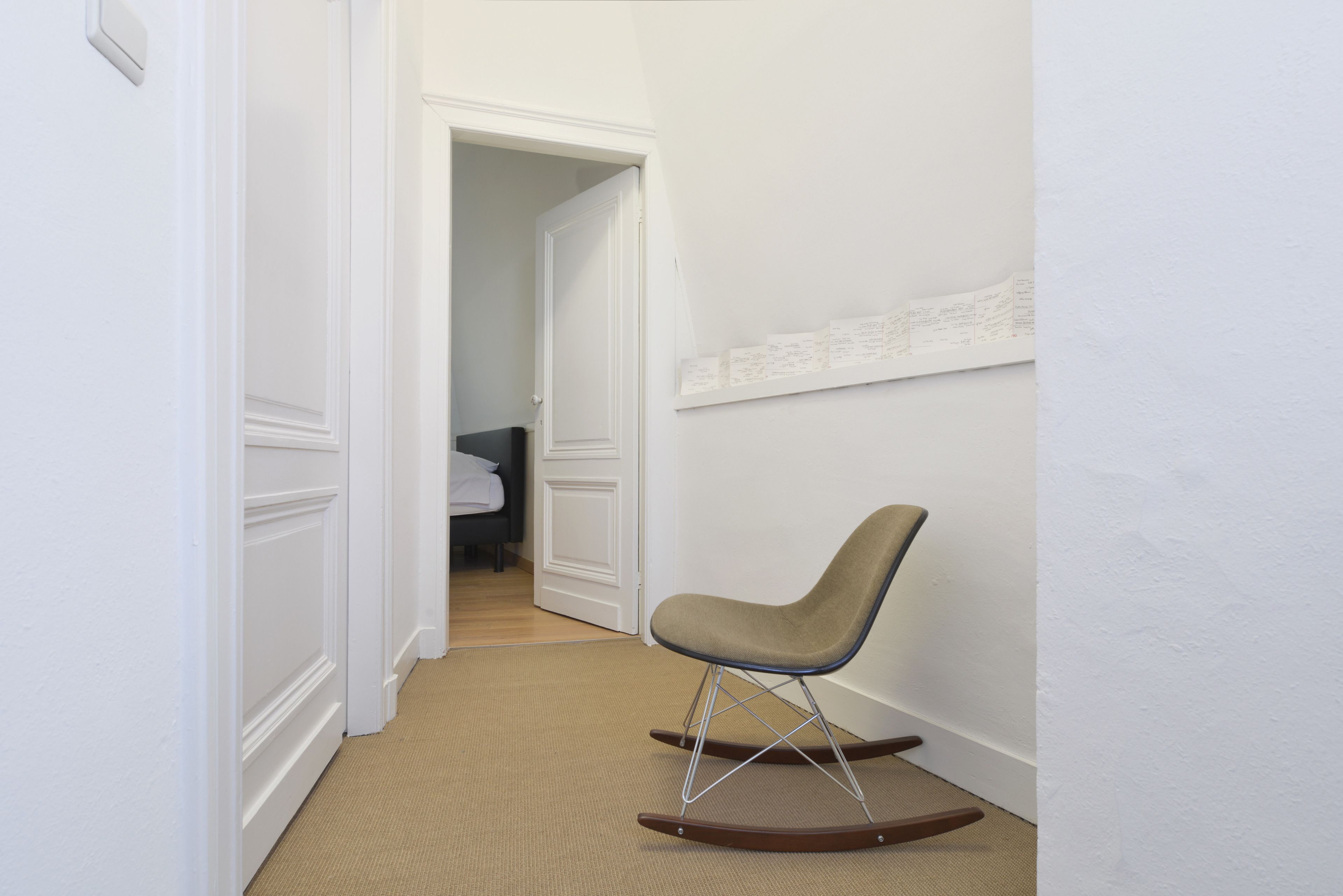 A narrow hallway with a rocking chair, a door leading to another room, and a wall-mounted shelf with papers.