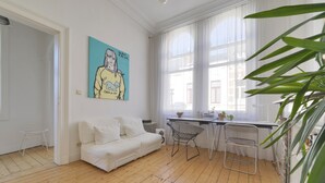 A room with a white sofa, a desk with a chair, a window with sheer curtains, and a painting on the wall. - Zurenborg Area Apartments (Antwerp)