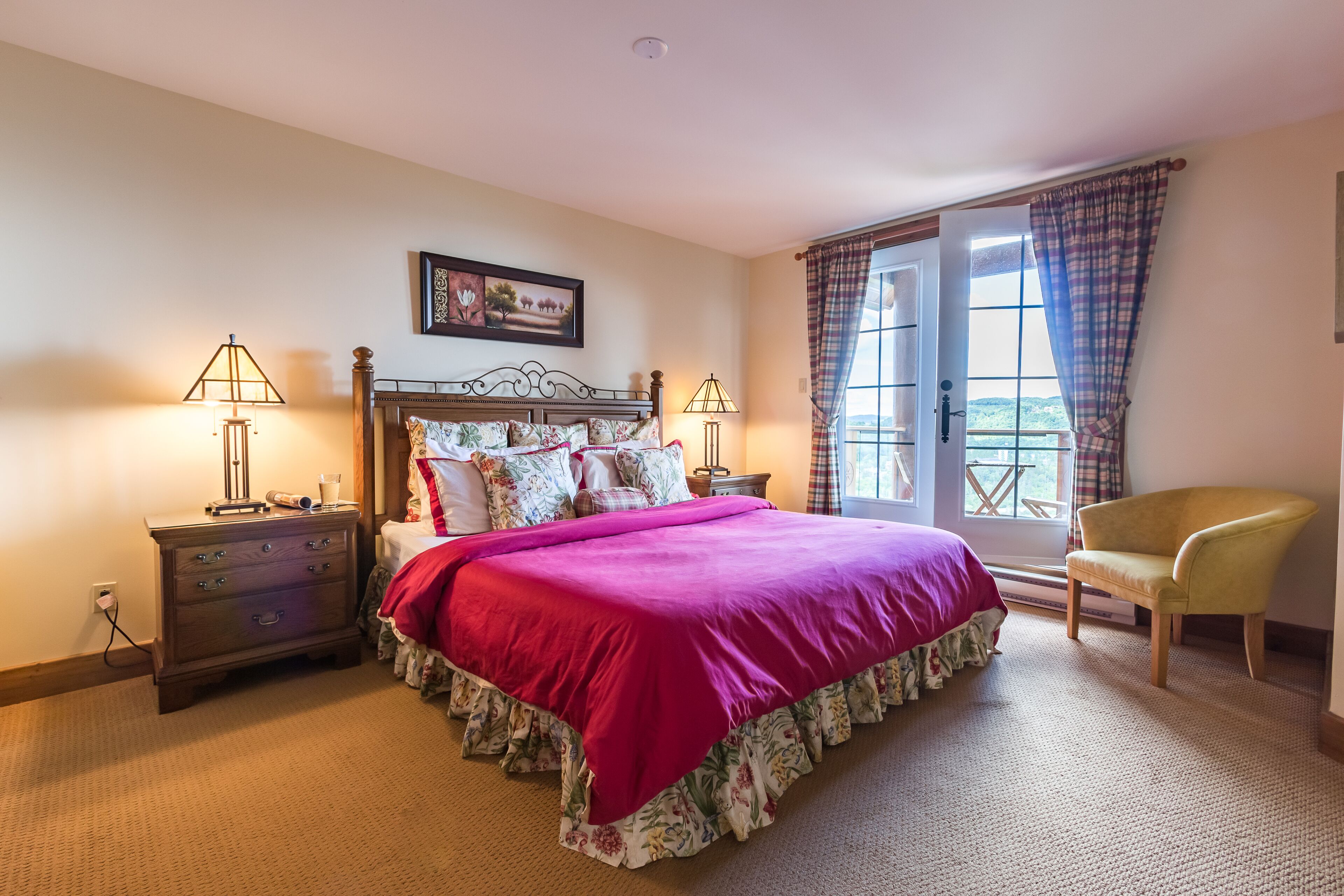 A bedroom with a large bed, two bedside tables with lamps, a chair, a window with curtains, and a painting on the wall.