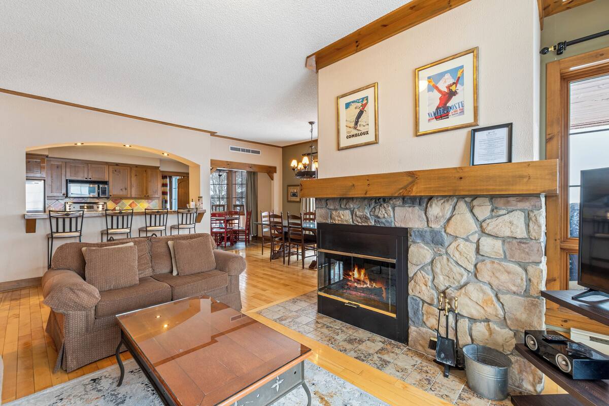 A spacious living room with a stone fireplace, a sofa, and a coffee table. There is a kitchen in the background with wooden cabinets and a dining area with chairs.