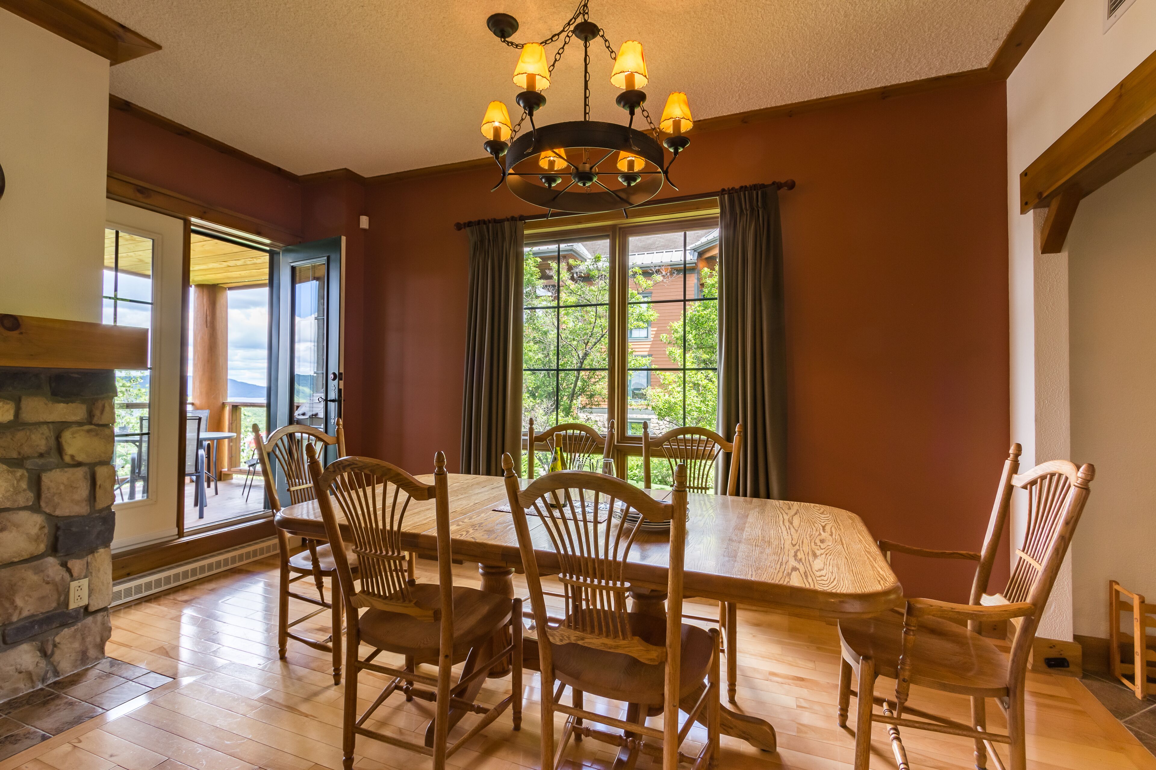 A dining area with a large wooden table and chairs, a chandelier, and a view through large windows.