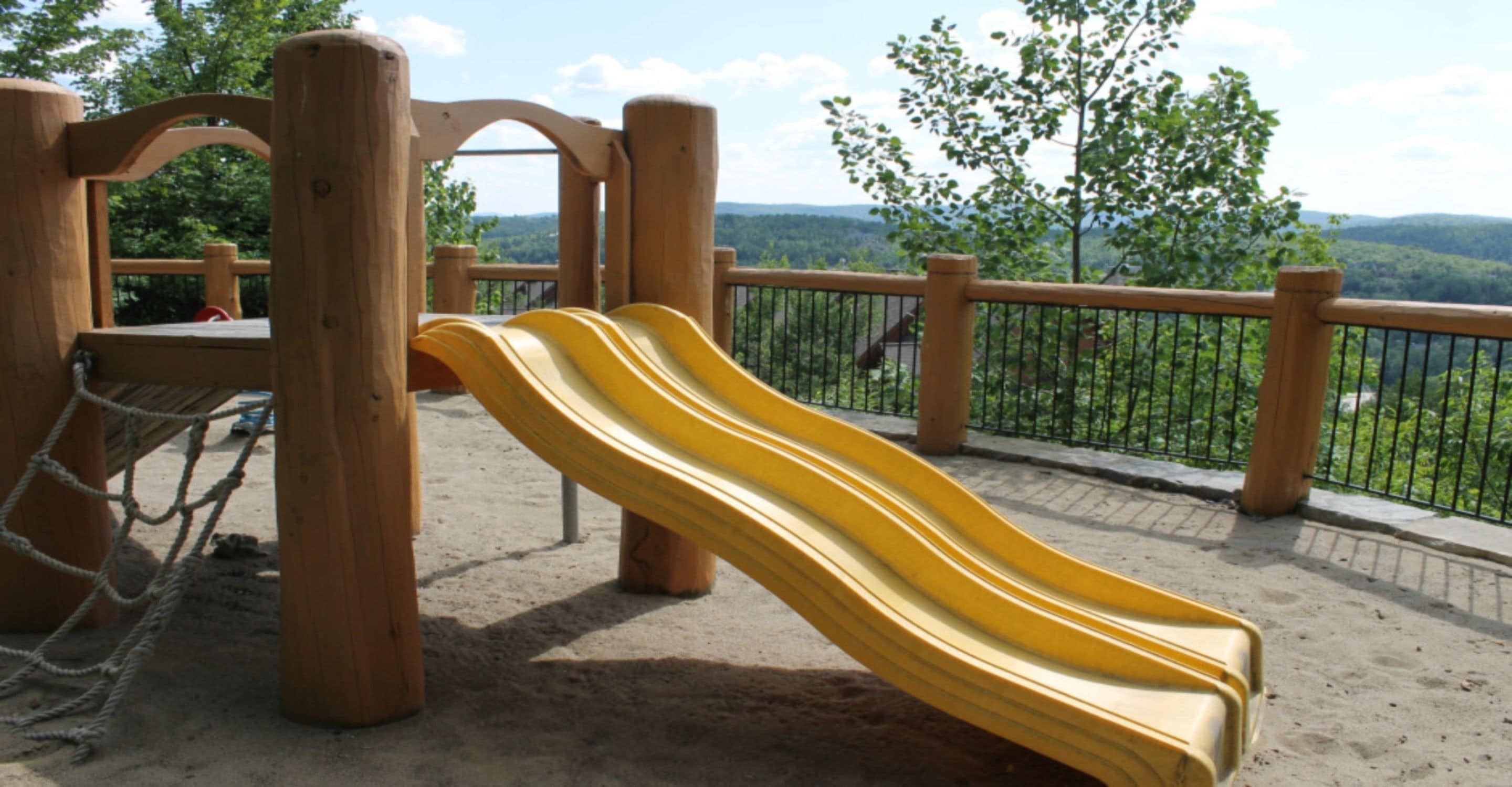 A playground with a yellow slide and a rope net, set against a backdrop of trees and hills.