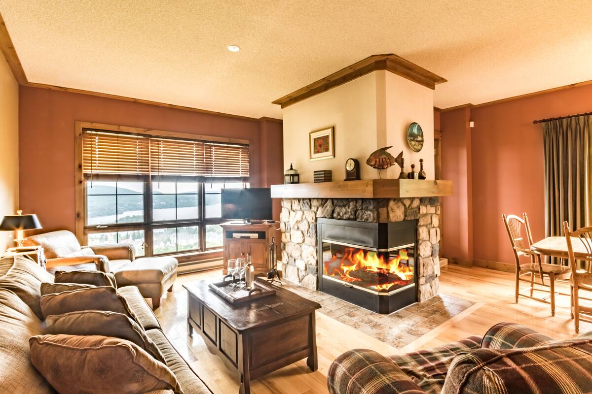 A cozy living room with a stone fireplace, a large window with blinds, a sofa, a coffee table, and a dining area with chairs.