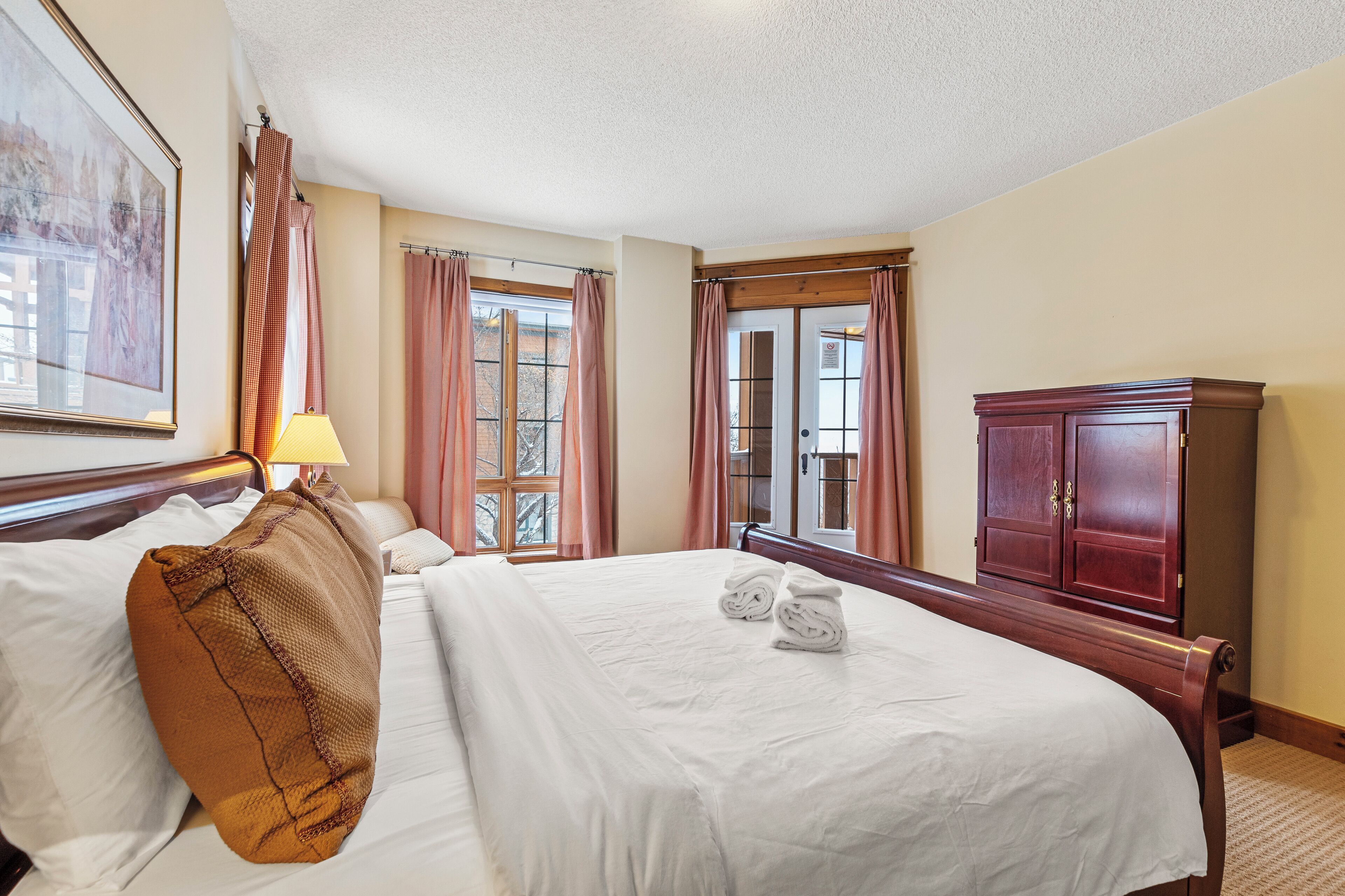 A hotel room with a large bed, a wooden wardrobe, a framed picture on the wall, and a window with curtains.