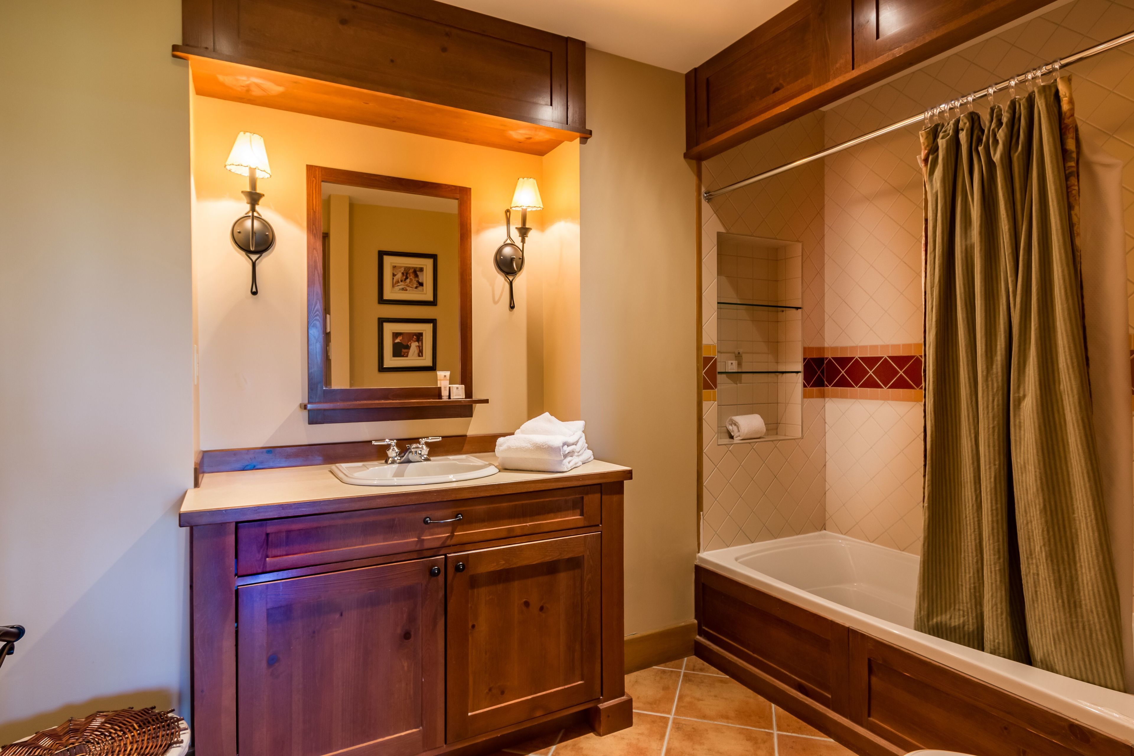 A bathroom with wooden cabinetry, a sink, a mirror with wall sconces, a shower with a curtain, and a tiled floor.