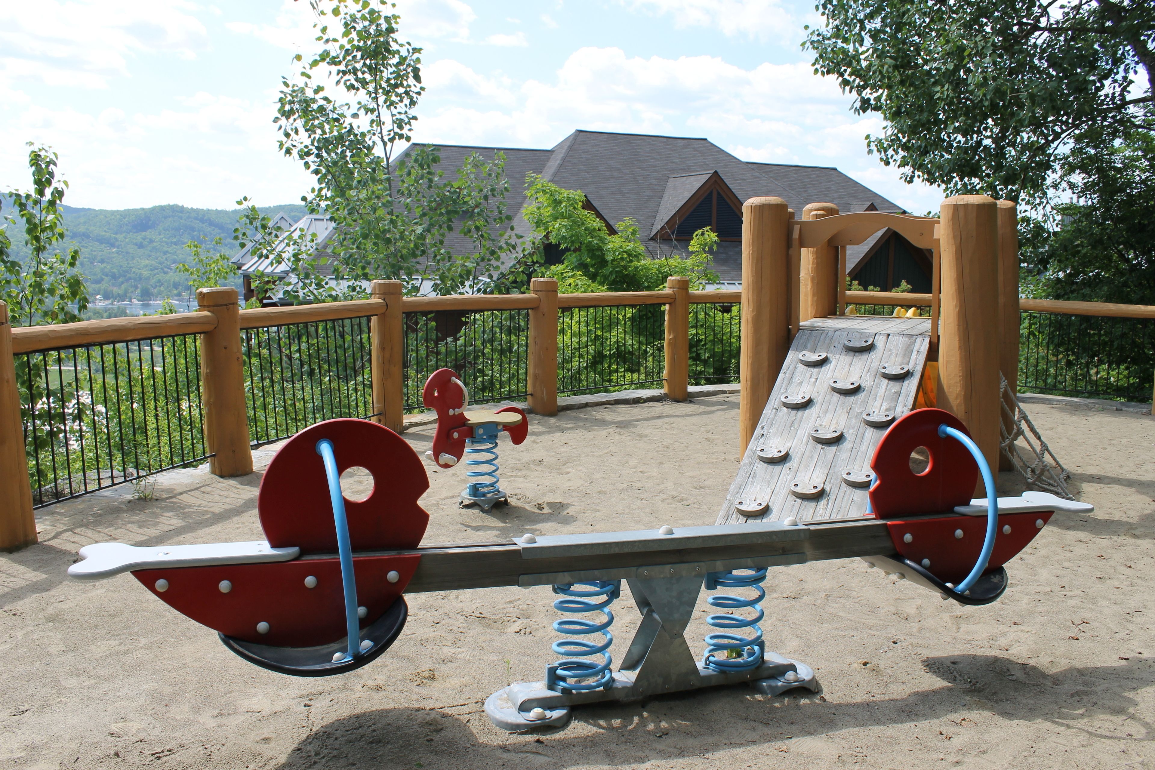 A playground with seesaw and swings, surrounded by a wooden fence and trees.