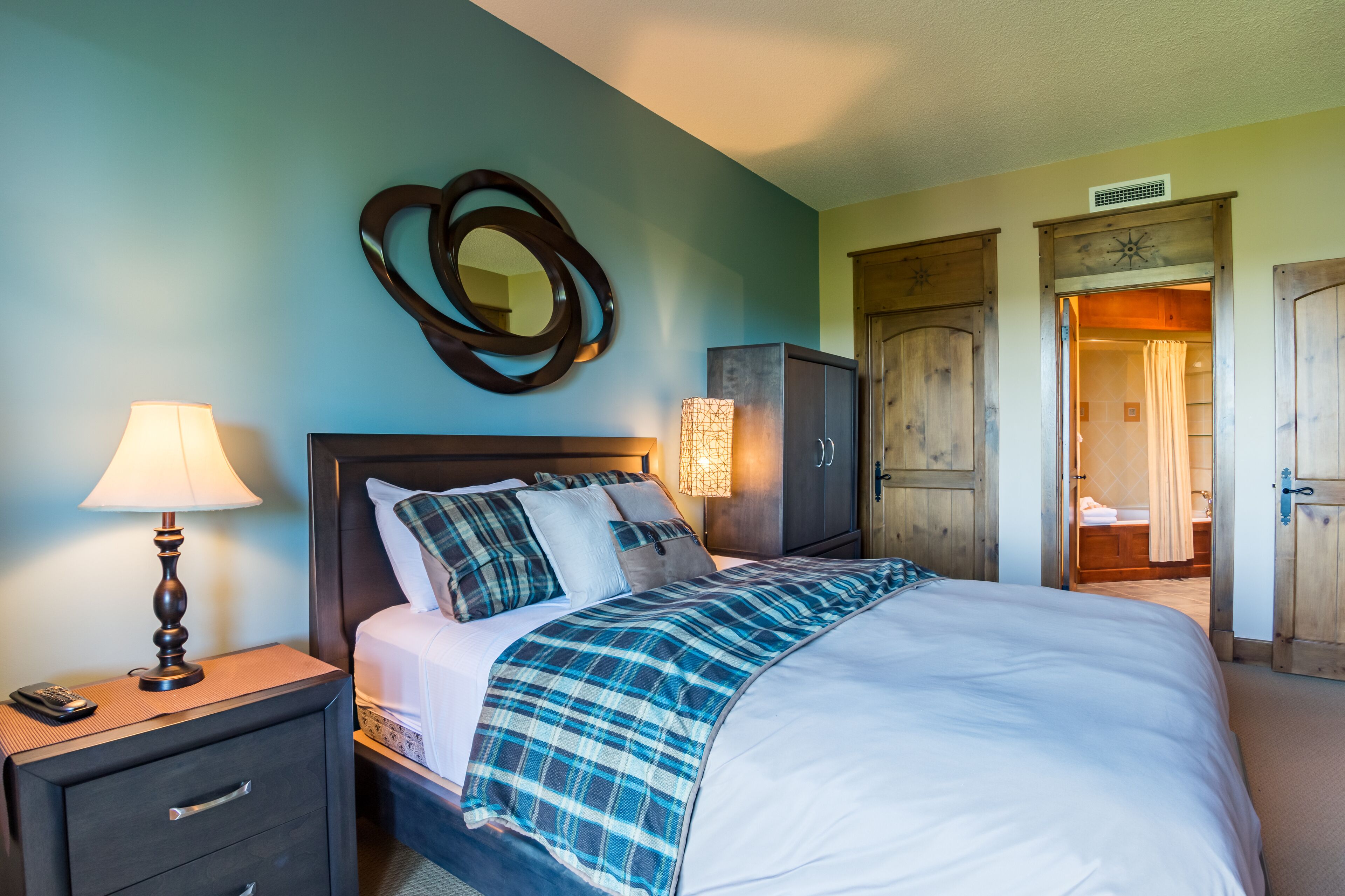 A bedroom with a bed, a nightstand with a lamp, a mirror, and a wooden wardrobe.