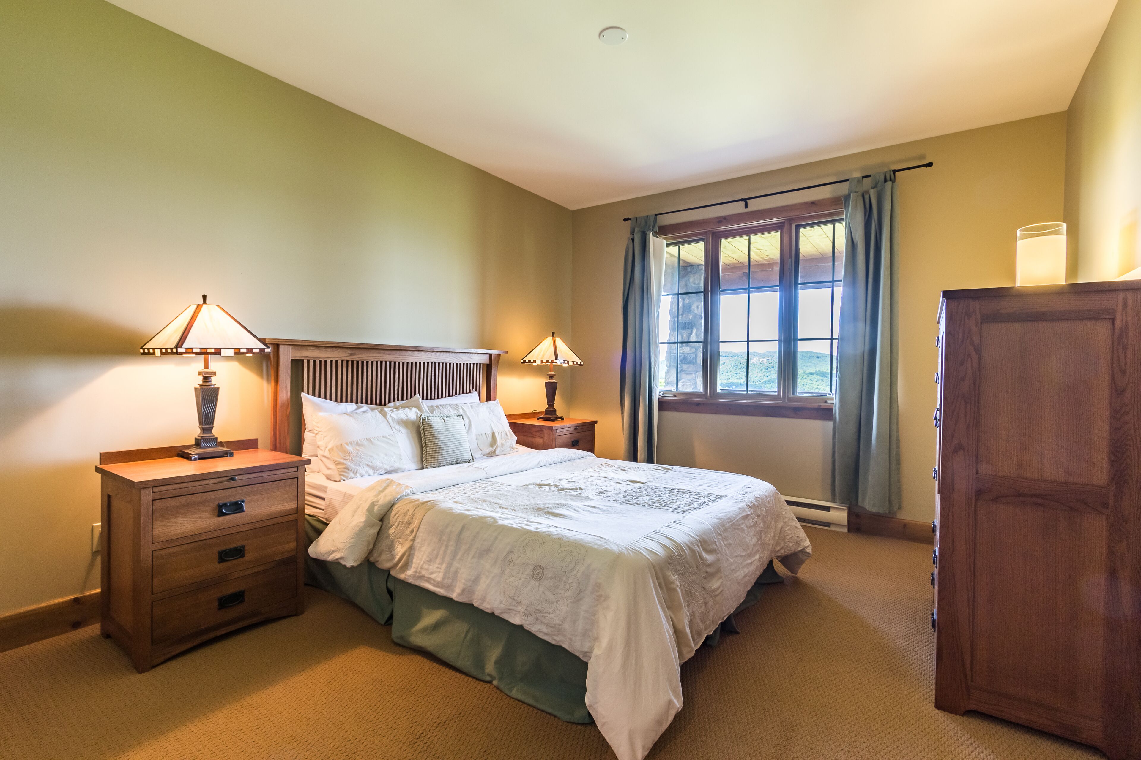 A bedroom with a bed, two bedside tables with lamps, a wooden dresser, and a window with curtains.