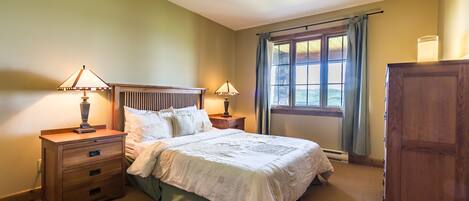 A bedroom with a bed, two bedside tables with lamps, a wooden dresser, and a window with curtains.