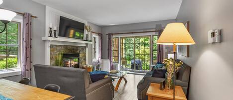 A modern living room with a fireplace, comfortable seating, a dining area, and large windows offering a view of greenery.