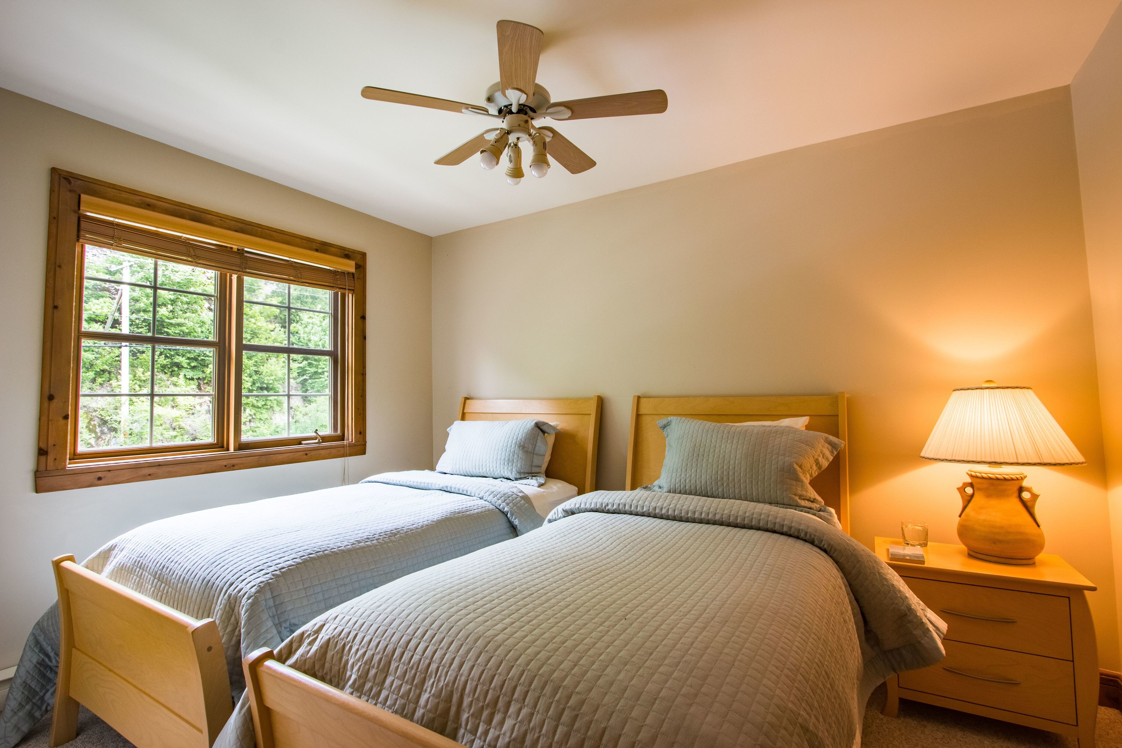 Ein Schlafzimmer mit zwei Betten, einem Deckenventilator, einem Fenster mit Jalousien und einem Nachttisch mit Lampe.