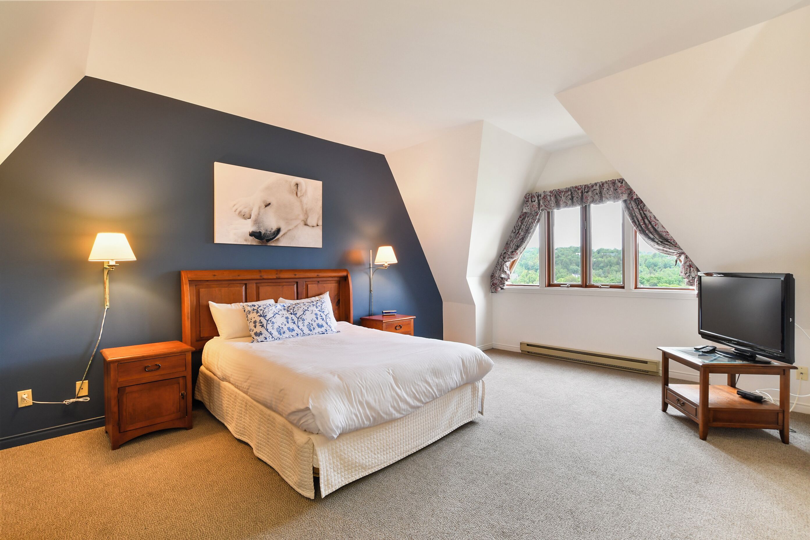 A bedroom with a bed, two bedside lamps, a TV on a stand, a painting on the wall, and a window with curtains.
