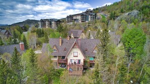 A large, multi-story house with a prominent balcony, surrounded by dense forest and hills in the background. - Le Plateau - RVMT (Mont-Tremblant)